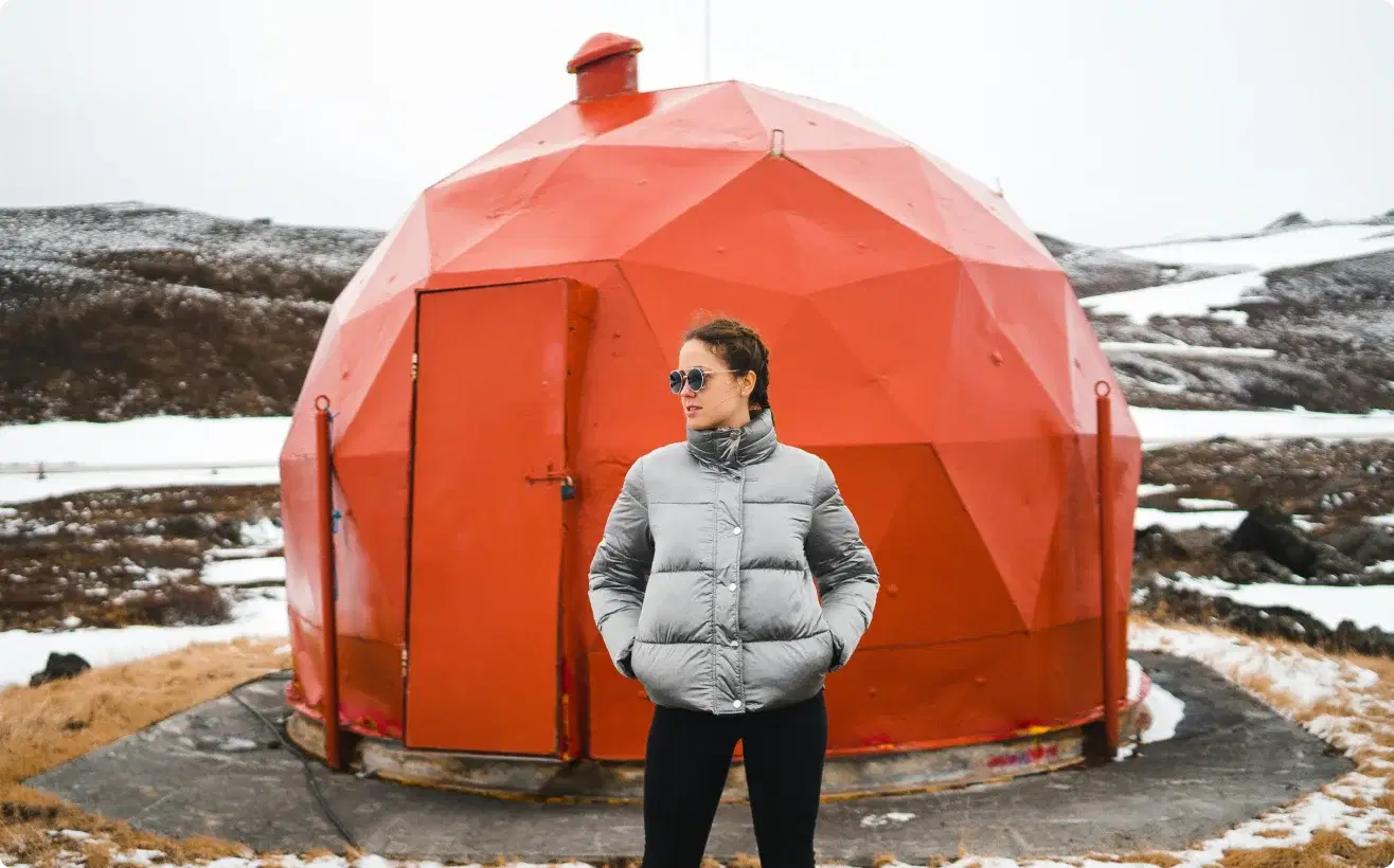 Persona con giacca invernale in piedi davanti a una struttura geodetica rossa unica in Islanda, simbolo di avventura e dell'opportunità di unirsi al programma di affiliazione di Cars Iceland.