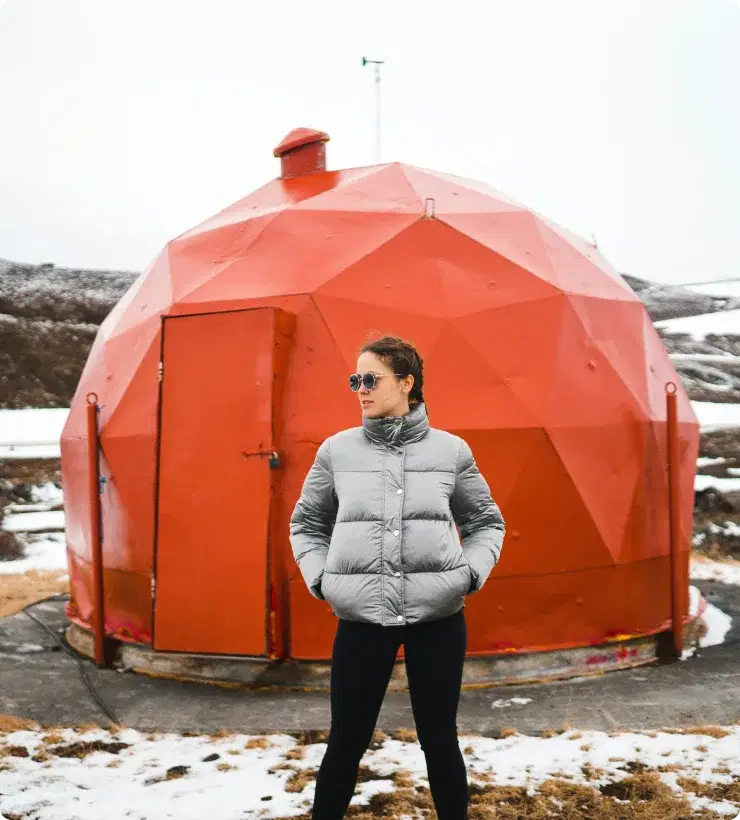 Persona con giacca invernale in piedi davanti a una struttura geodetica rossa unica in Islanda, simbolo di avventura e dell'opportunità di unirsi al programma di affiliazione di Cars Iceland.