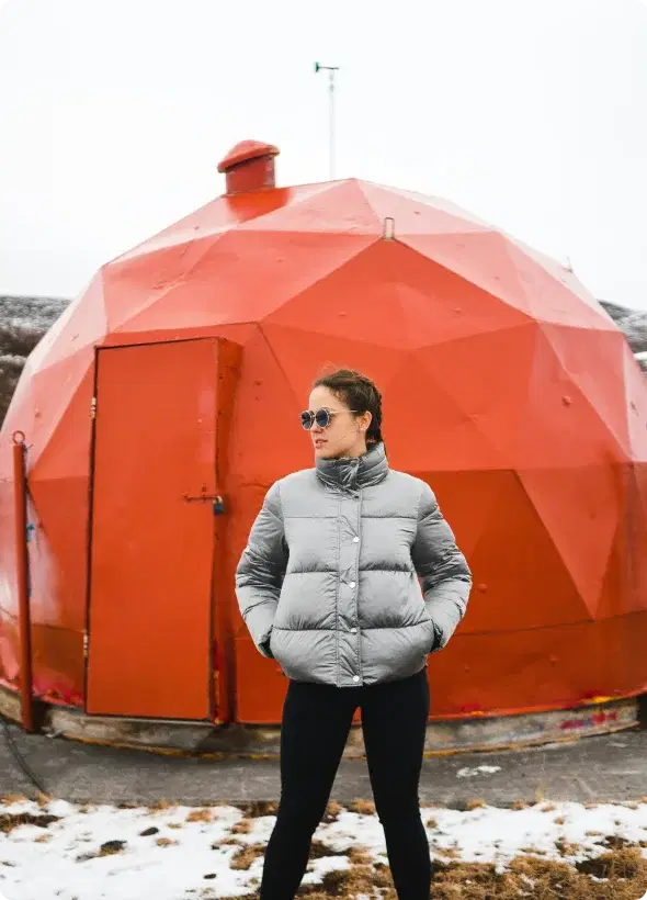 Persona con giacca invernale in piedi davanti a una struttura geodetica rossa unica in Islanda, simbolo di avventura e dell'opportunità di unirsi al programma di affiliazione di Cars Iceland.