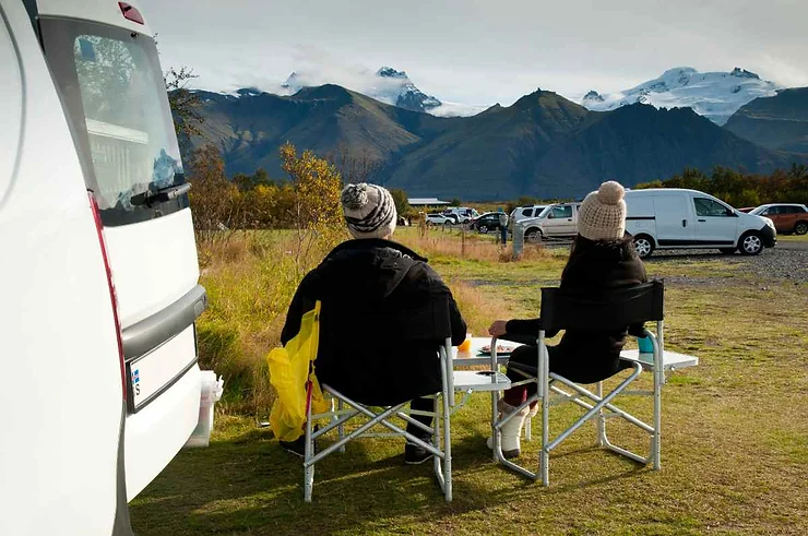Due persone sedute su sedie pieghevoli accanto a un camper, con vista sulle montagne innevate in Islanda.