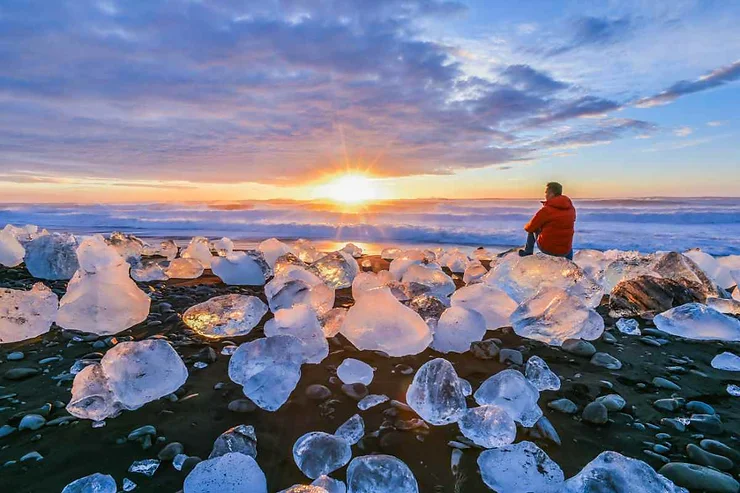 Scogliere di ghiaccio sulla spiaggia nera islandese al tramonto, con una persona in giacca rossa che ammira l'oceano.