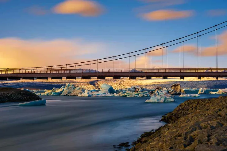 Ponte sospeso con blocchi di ghiaccio e fiume in primo piano, sotto un cielo colorato al tramonto in Islanda.