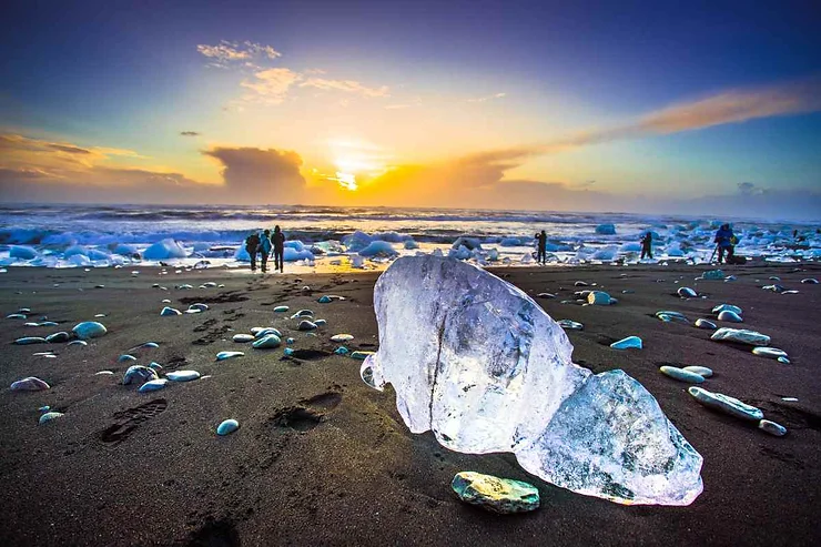 Tramonto sulla spiaggia dei diamanti in Islanda, con un grande blocco di ghiaccio trasparente in primo piano e l'oceano sullo sfondo.