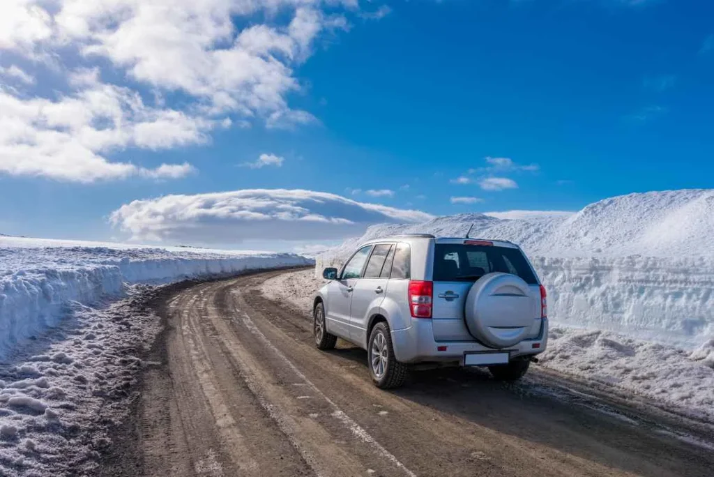 Guidare nelle condizioni invernali islandesi Un SUV bianco che guida su una strada sgomberata dalla neve circondata da alti cumuli di neve sotto un cielo blu brillante in Islanda.