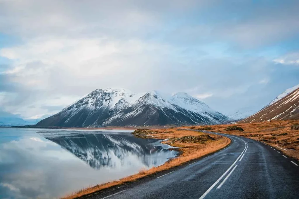 Strade asfaltate in Islanda: percorso panoramico con vista sulle montagne Una strada asfaltata panoramica accanto a un lago calmo con montagne innevate riflesse sull'acqua sotto un cielo pastello.
