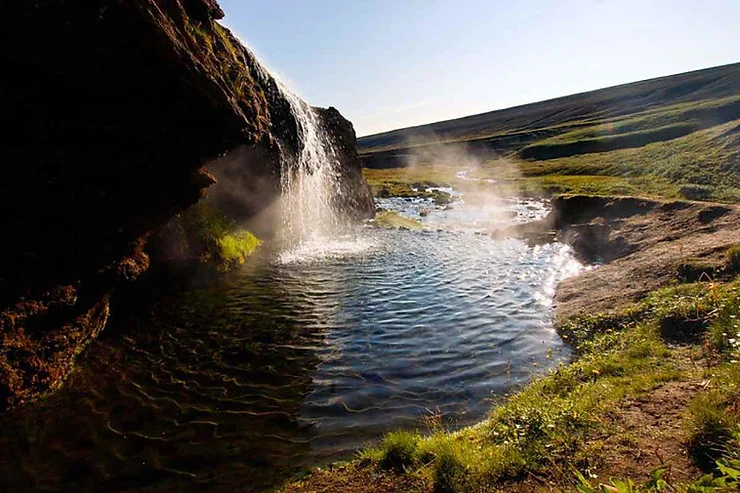 Una cascata termale scende da una roccia muschiosa in una piscina naturale, circondata da un paesaggio verde e rilassante.