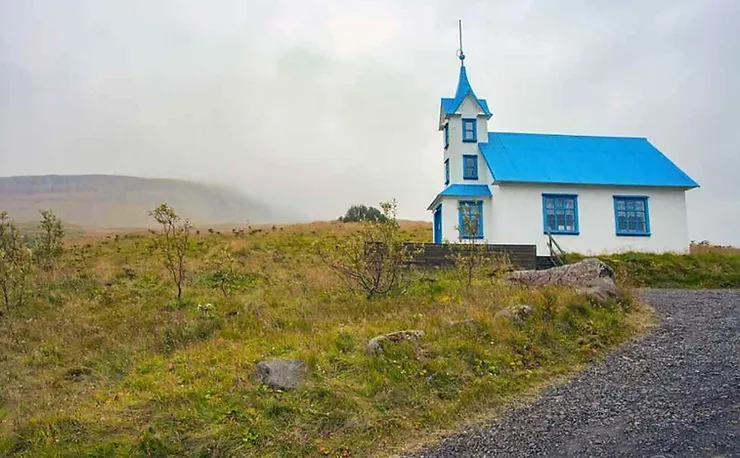 Una piccola chiesa bianca con tetto blu sorge solitaria in una campagna islandese nebbiosa, creando un’atmosfera tranquilla e misteriosa.