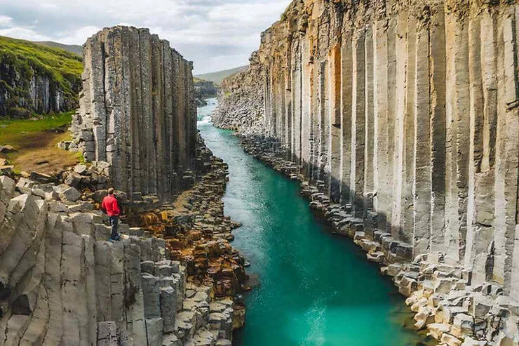 La gola di Stuðlagil con colonne basaltiche perfette e un fiume turchese brillante, dove un escursionista ammira il paesaggio mozzafiato.
