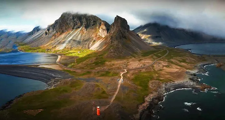 Veduta aerea di una spettacolare penisola islandese con montagne scoscese, spiagge nere e un piccolo faro rosso che si staglia sul paesaggio.