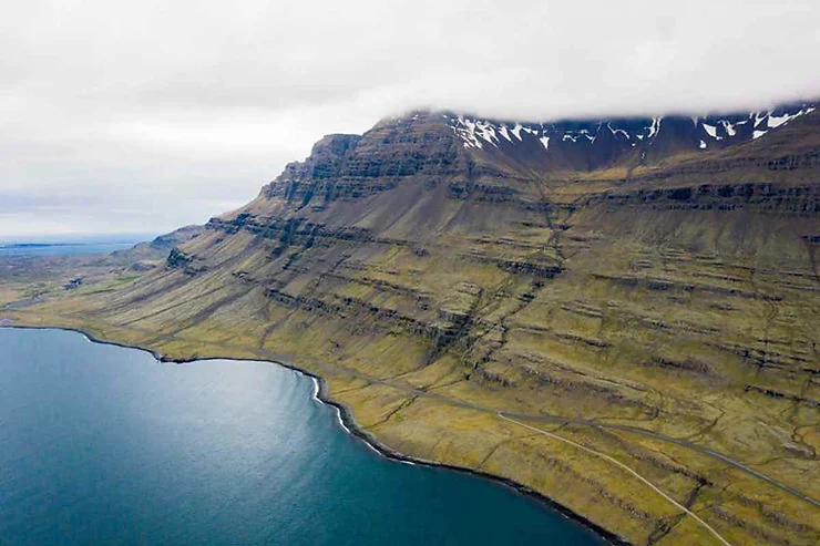 Veduta aerea di una maestosa montagna islandese avvolta da nuvole basse, con pendii verdi che incontrano le acque calme dell'oceano.