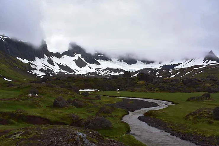 Un ruscello sinuoso attraversa una vallata verdeggiante con montagne innevate e nebbiose sullo sfondo in Islanda.