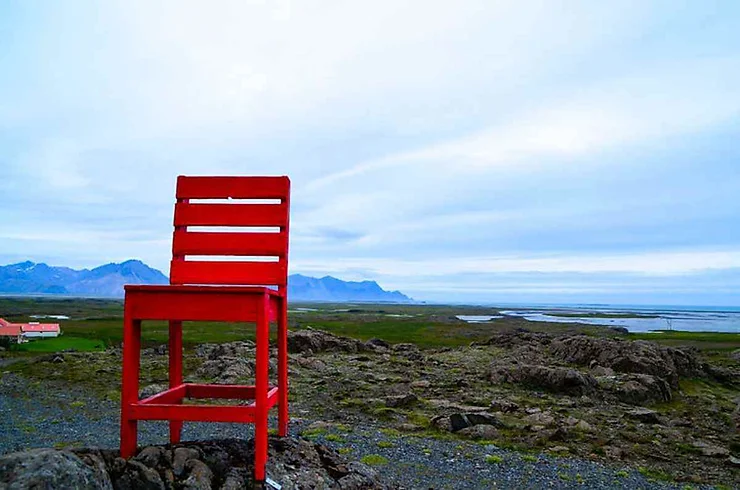 Una grande sedia rossa solitaria su una collina rocciosa con vista su un vasto paesaggio islandese e l'oceano all'orizzonte.