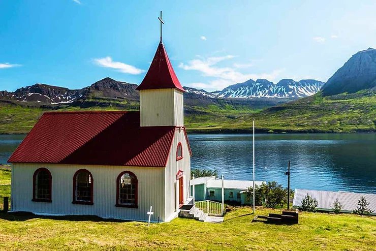 Una piccola chiesa islandese bianca con tetto rosso, situata accanto a un lago calmo con montagne innevate sullo sfondo.