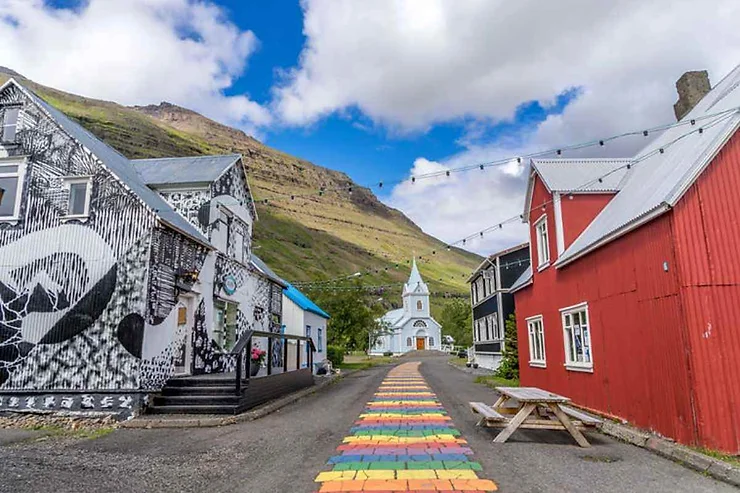Una strada colorata e vivace in un villaggio islandese, con edifici tradizionali e una chiesa bianca incorniciata dalle montagne.
