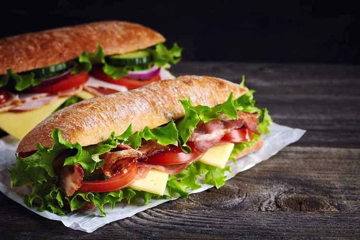 Due panini freschi con insalata, pomodori, formaggio e pancetta su carta bianca.