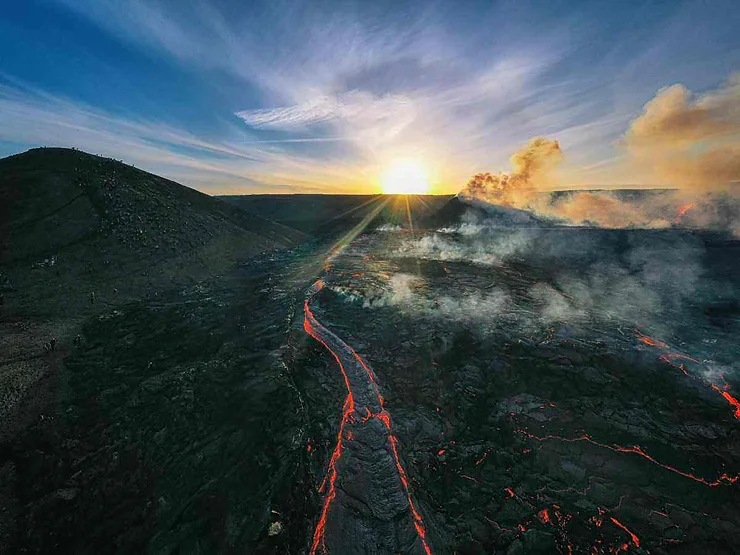 Colata di lava su un vulcano islandese al tramonto, con cielo sereno e fumo in aumento.