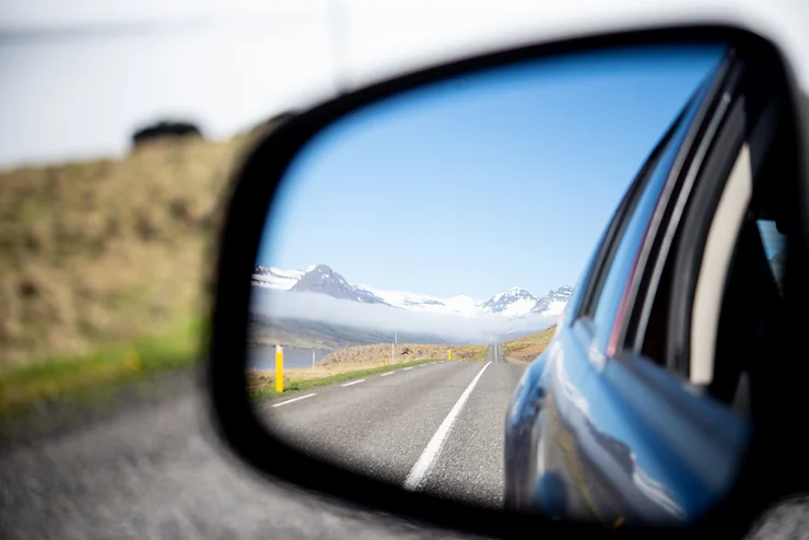 Una strada islandese che si estende fino alle montagne innevate, vista attraverso lo specchietto retrovisore di un'auto.