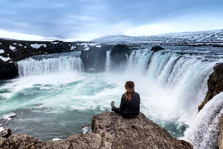 Una spettacolare cascata a ferro di cavallo, con un viaggiatore seduto su una roccia che osserva il flusso potente dell'acqua. Sullo sfondo si vedono montagne innevate.