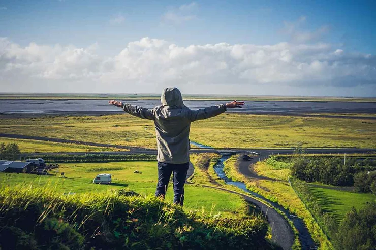 Una persona con le braccia aperte su una collina verde che si affaccia su un paesaggio islandese ampio e pianeggiante.