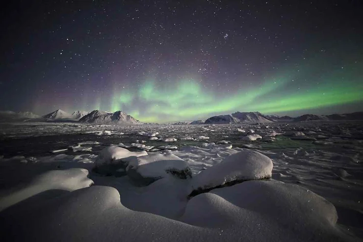 Un paesaggio innevato illuminato dall'aurora boreale con un cielo stellato sopra ghiacciai e montagne.