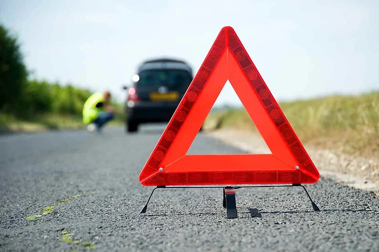 Un triangolo di emergenza rosso brillante è posizionato su una strada asfaltata. Sullo sfondo, una persona si avvicina a un'auto ferma sul ciglio della strada. La scena rappresenta una situazione di emergenza o guasto stradale.