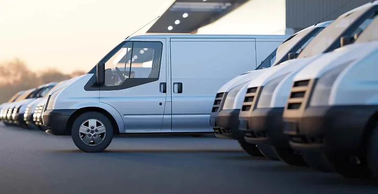 Una fila di furgoni bianchi è parcheggiata ordinatamente, probabilmente in un'area di noleggio o deposito. La scena è ambientata al tramonto, con una luce calda che si riflette sui veicoli.