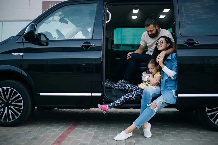 Una famiglia è seduta accanto a un minivan nero. Una bambina si appoggia alla madre, mentre il padre è accovacciato accanto a loro. Sembrano rilassati e felici, pronti per un viaggio.