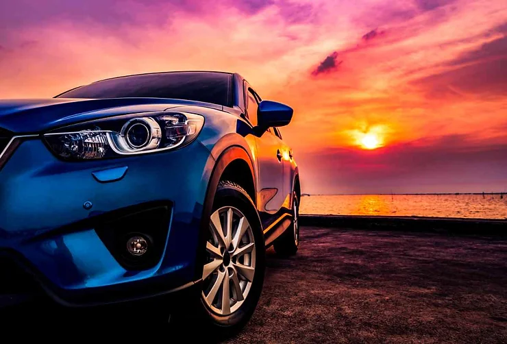  Una macchina blu metallizzata è parcheggiata su una strada vicino a un lago. Sullo sfondo si vede un tramonto spettacolare con sfumature rosa, arancioni e viola.