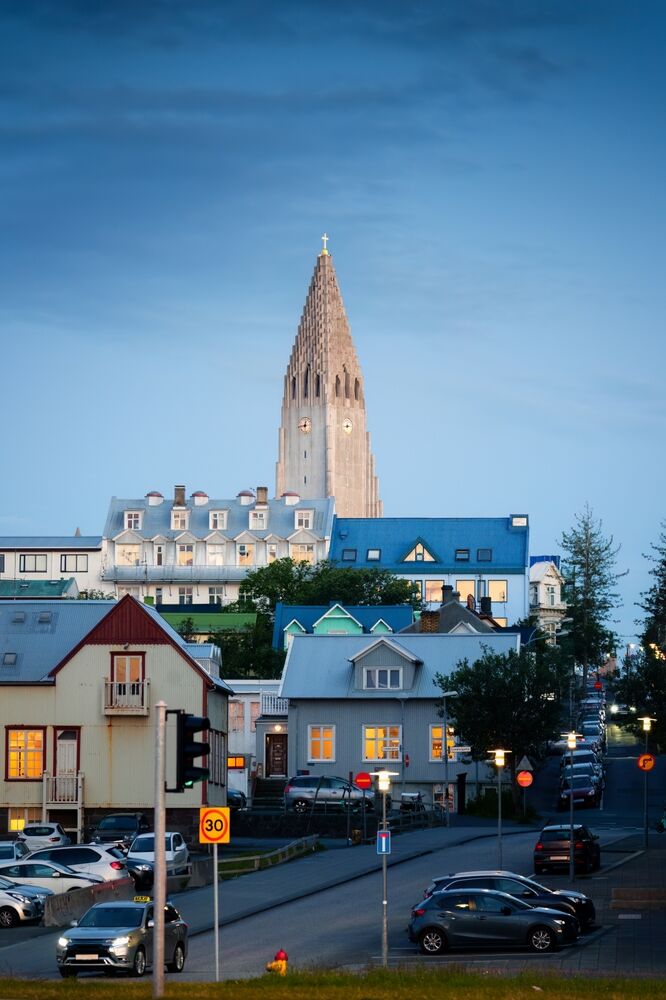 Vista del centro di Reykjavik con una strada illuminata al crepuscolo e la chiesa Hallgrímskirkja che svetta sullo sfondo. Case colorate e auto parcheggiate dominano il paesaggio.