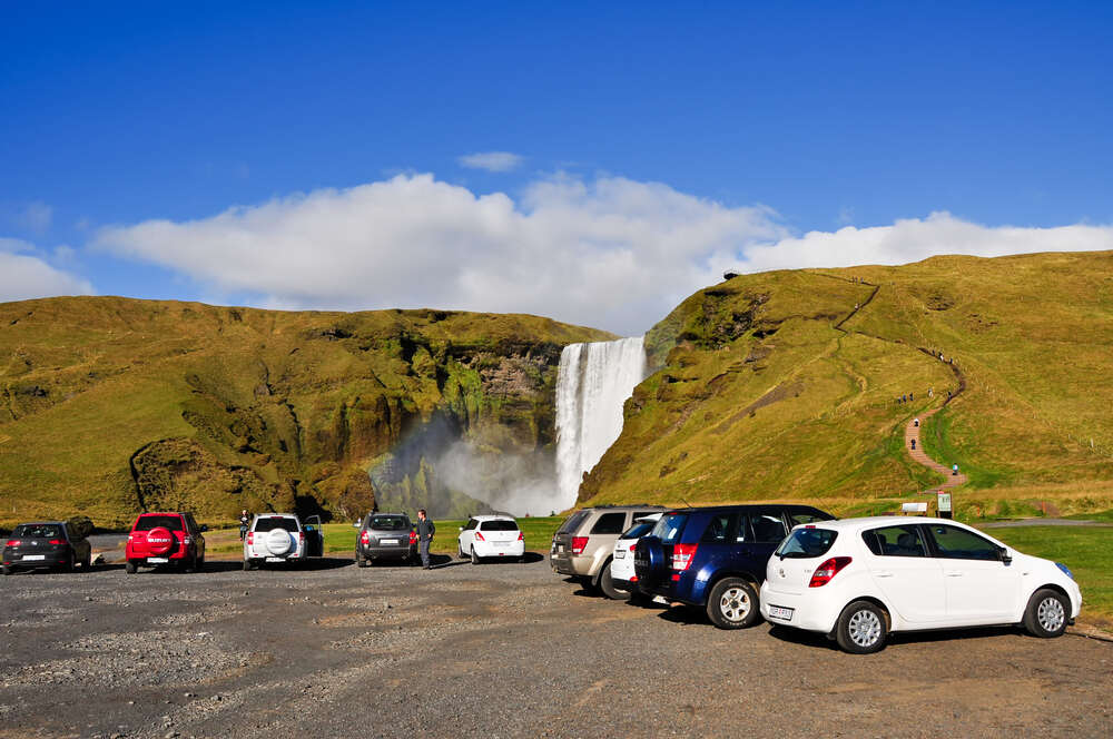 Un parcheggio all'aperto con diverse auto parcheggiate, situato di fronte a una magnifica cascata circondata da colline verdi sotto un cielo blu.