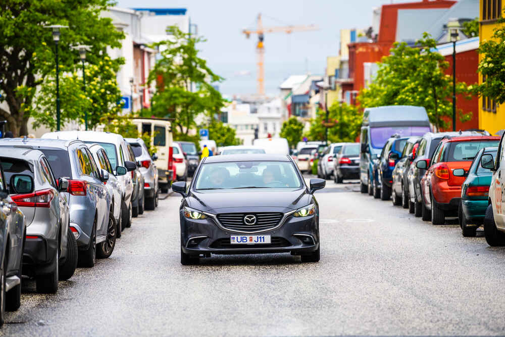 Una Mazda nera guida su una strada cittadina affiancata da auto parcheggiate. Gli alberi verdi e gli edifici colorati creano un'atmosfera vivace.