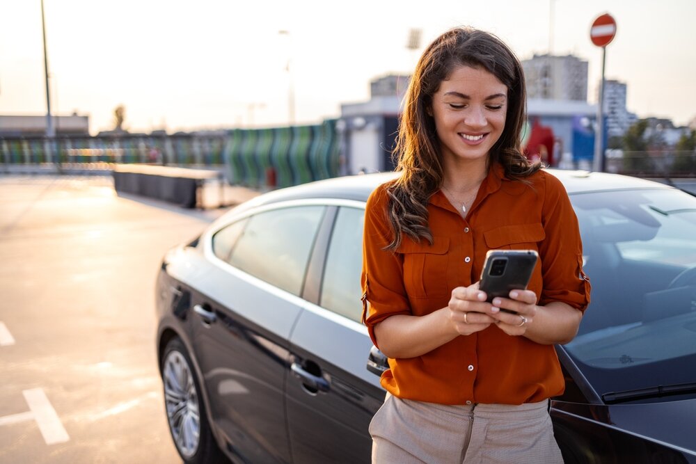 Una donna sorridente con una camicia arancione utilizza il cellulare appoggiata a un'auto moderna in un parcheggio sotto il sole.