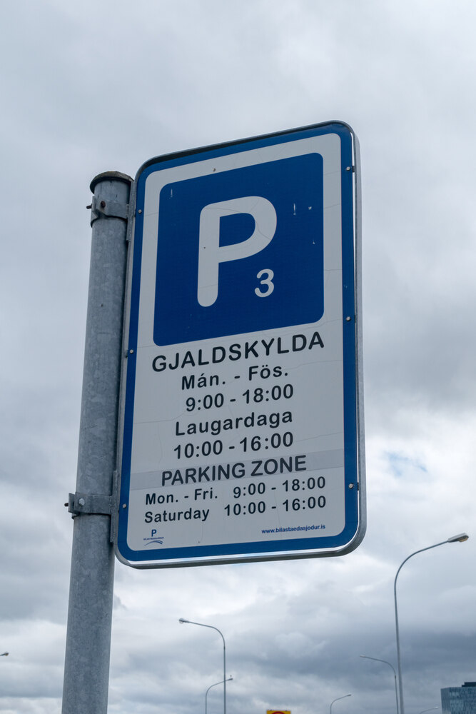 Un cartello di parcheggio blu e bianco che indica gli orari e le regole per il parcheggio a Reykjavik, scritto in islandese e in inglese, su uno sfondo di cielo nuvoloso.