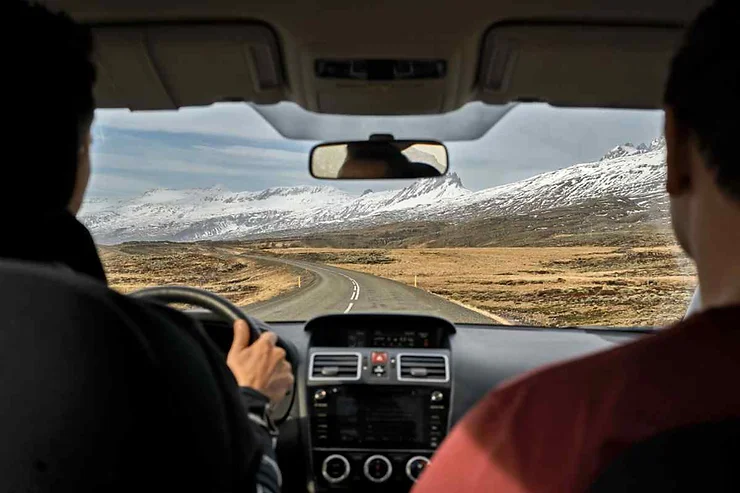 Vista dall'interno di un'auto in movimento con due persone a bordo, che percorre una strada verso un paesaggio con montagne innevate.