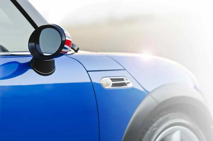 Dettaglio della carrozzeria di un'auto blu con specchietto laterale decorato con una bandiera britannica. La luce solare si riflette delicatamente sulla superficie lucida.