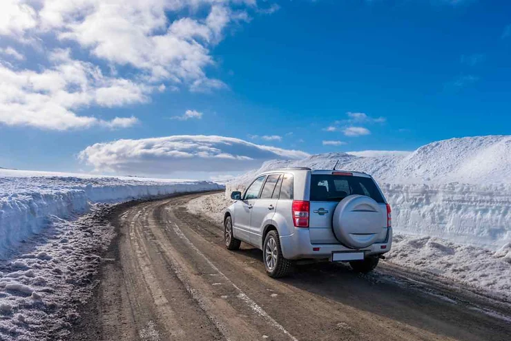 Un SUV grigio argento percorre una strada sterrata circondata da cumuli di neve alti sotto un cielo blu con nuvole soffici.