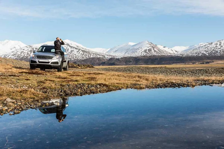 Un SUV grigio parcheggiato vicino a uno specchio d'acqua con montagne innevate sullo sfondo. Una persona sta scattando foto accanto al veicolo.