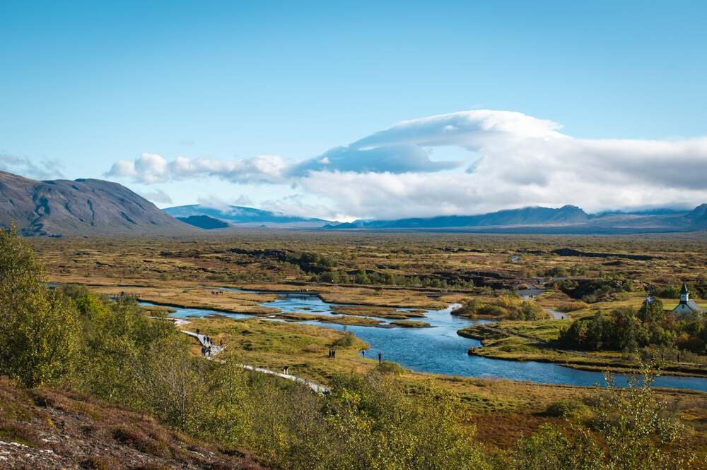 Un paesaggio mozzafiato nel Parco Nazionale di Thingvellir, con un fiume che attraversa una vasta pianura circondata da montagne in lontananza sotto un cielo limpido e azzurro.