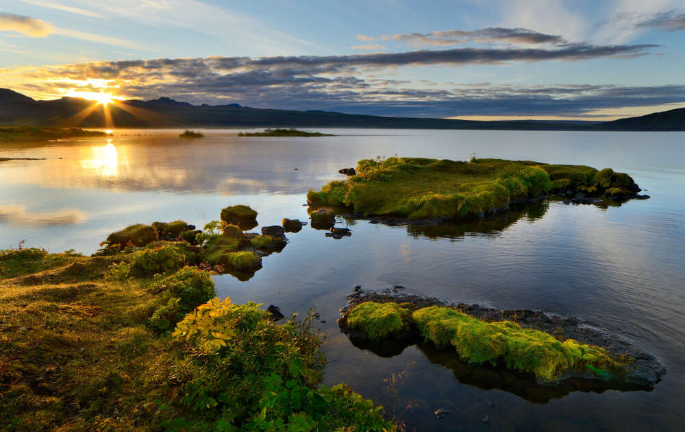 Uno scenario suggestivo al tramonto presso il lago Thingvallavatn, con il sole che si riflette sull'acqua calma, piccole isole coperte di muschio e un cielo parzialmente nuvoloso.