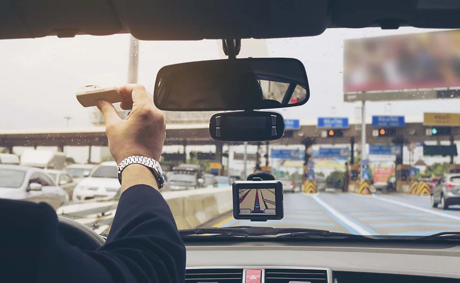 Un conducente all’interno di un’auto allunga la mano verso il parabrezza con un dispositivo per il pedaggio, pronto a entrare nella corsia del casello; sul cruscotto un GPS mostra la navigazione e si vedono altri veicoli davanti.