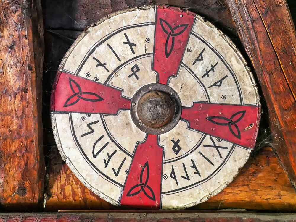 Un grande scudo circolare di legno con sezioni dipinte in rosso e bianco, decorato con rune e simboli nordici. Al centro è presente un bossolo metallico rotondo.