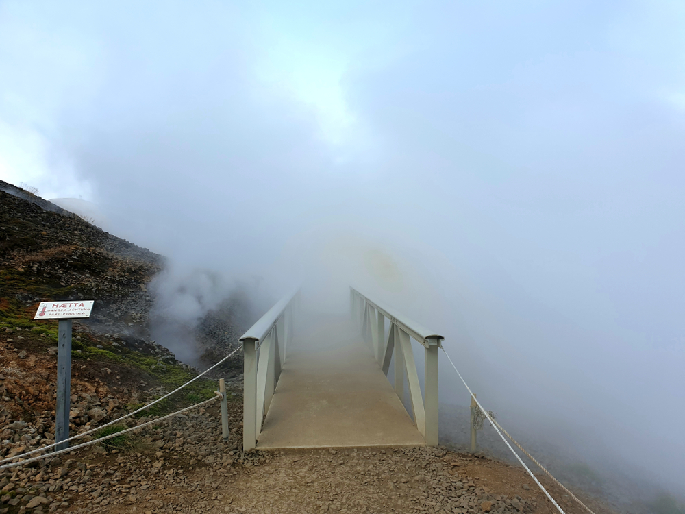 Ponte immerso nel vapore geotermico nell’area termale di Hverir, Islanda, con cartello di pericolo che avverte delle alte temperature.