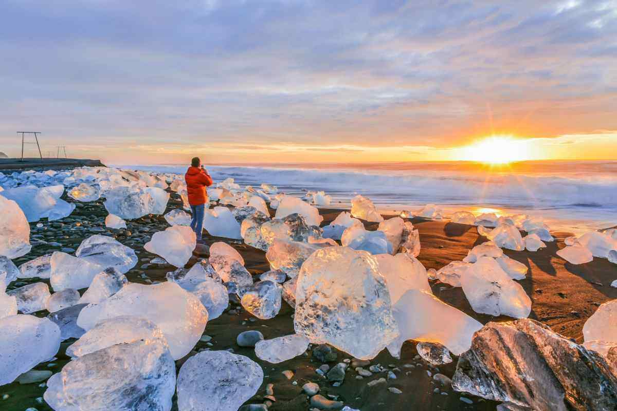 Diamond Beach in Islanda al tramonto, con blocchi di ghiaccio traslucidi sulla sabbia nera e un viaggiatore che fotografa il paesaggio artico.