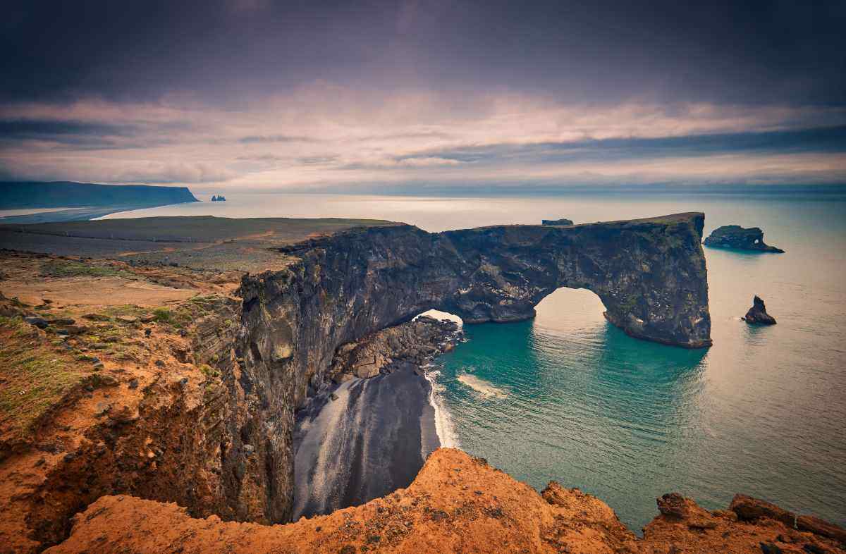 Vista aerea di Dyrhólaey in Islanda con l’arco naturale di lava nera affacciato sull’oceano e la spiaggia nera sottostante, al tramonto nuvoloso.