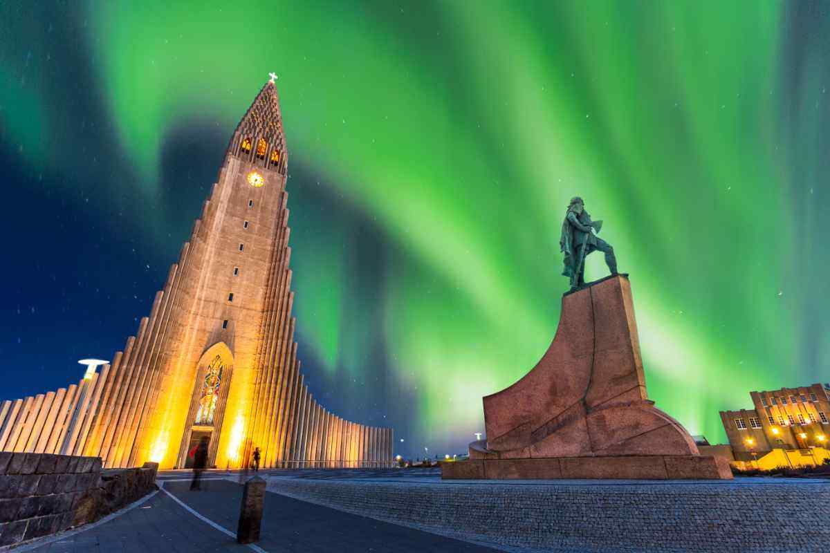 La chiesa Hallgrímskirkja illuminata a Reykjavík con aurora boreale verde nel cielo notturno, accanto alla statua di Leifur Eiríksson.