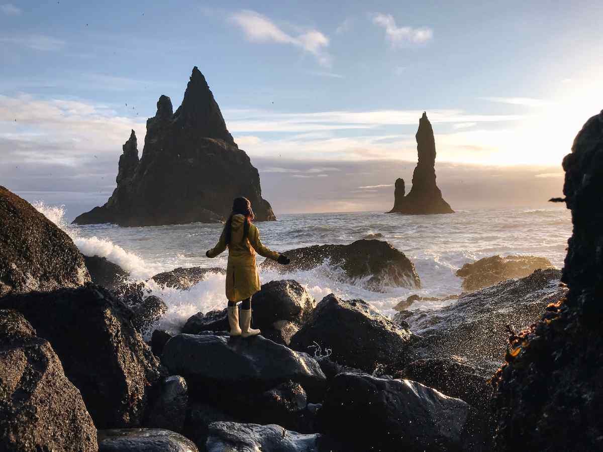 Viaggiatrice sulla spiaggia nera di Reynisfjara in Islanda osserva i faraglioni Reynisdrangar, tra onde impetuose e luce dorata del tramonto.