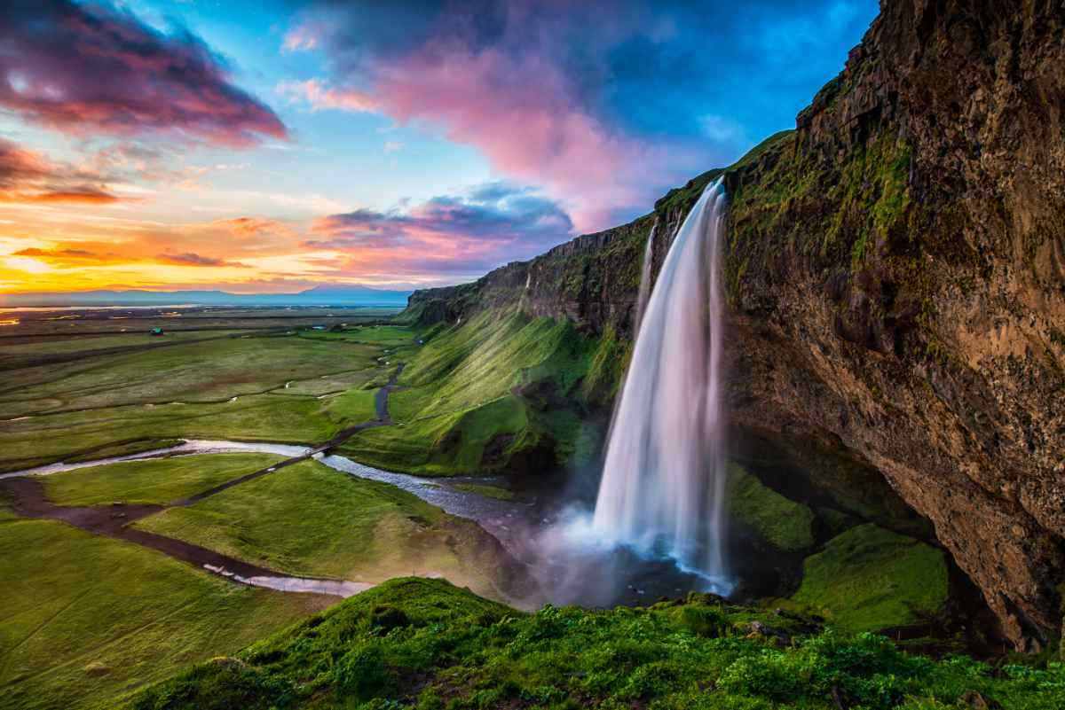 La cascata Seljalandsfoss in Islanda vista al tramonto, con paesaggi verdi e cielo colorato, famosa per il sentiero che passa dietro l’acqua.