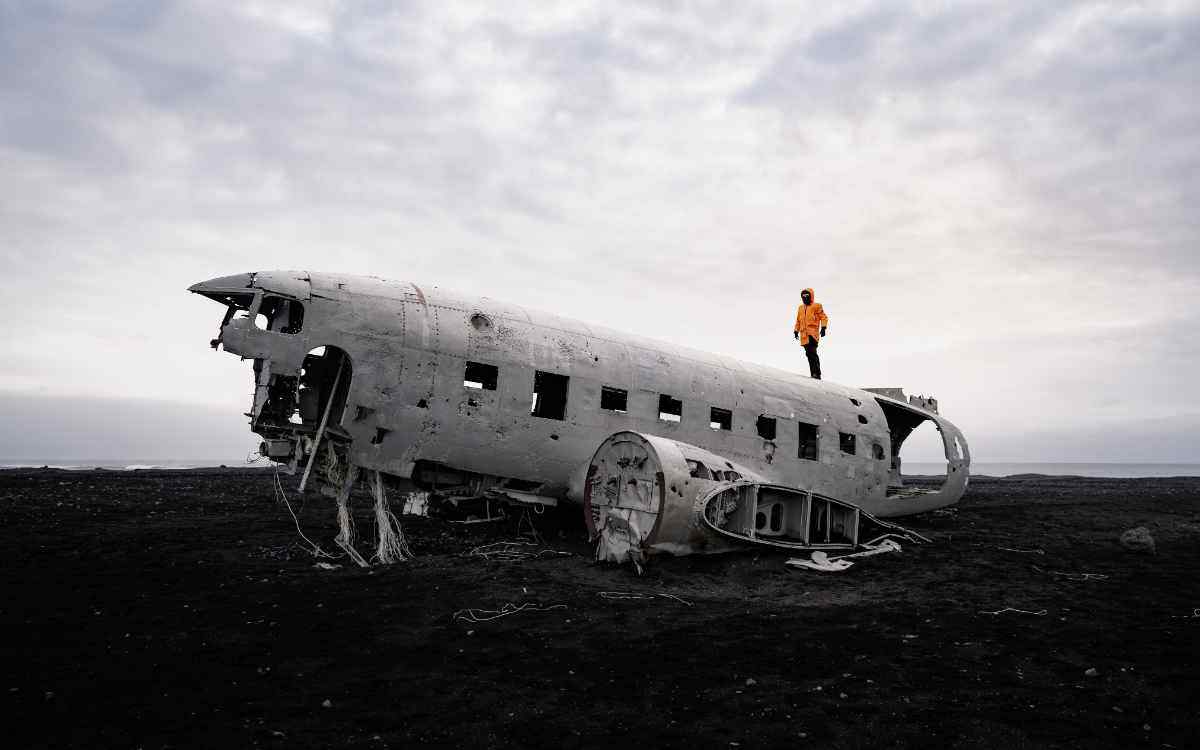 Relitto dell’aereo DC-3 sulla spiaggia nera di Sólheimasandur in Islanda, con viaggiatore in giacca arancione che osserva il paesaggio surreale.