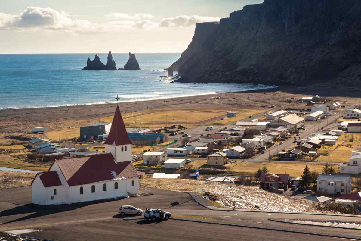 Vista panoramica di Vík í Mýrdal in Islanda con la chiesa dal tetto rosso e i faraglioni Reynisdrangar che emergono dall’oceano Atlantico.