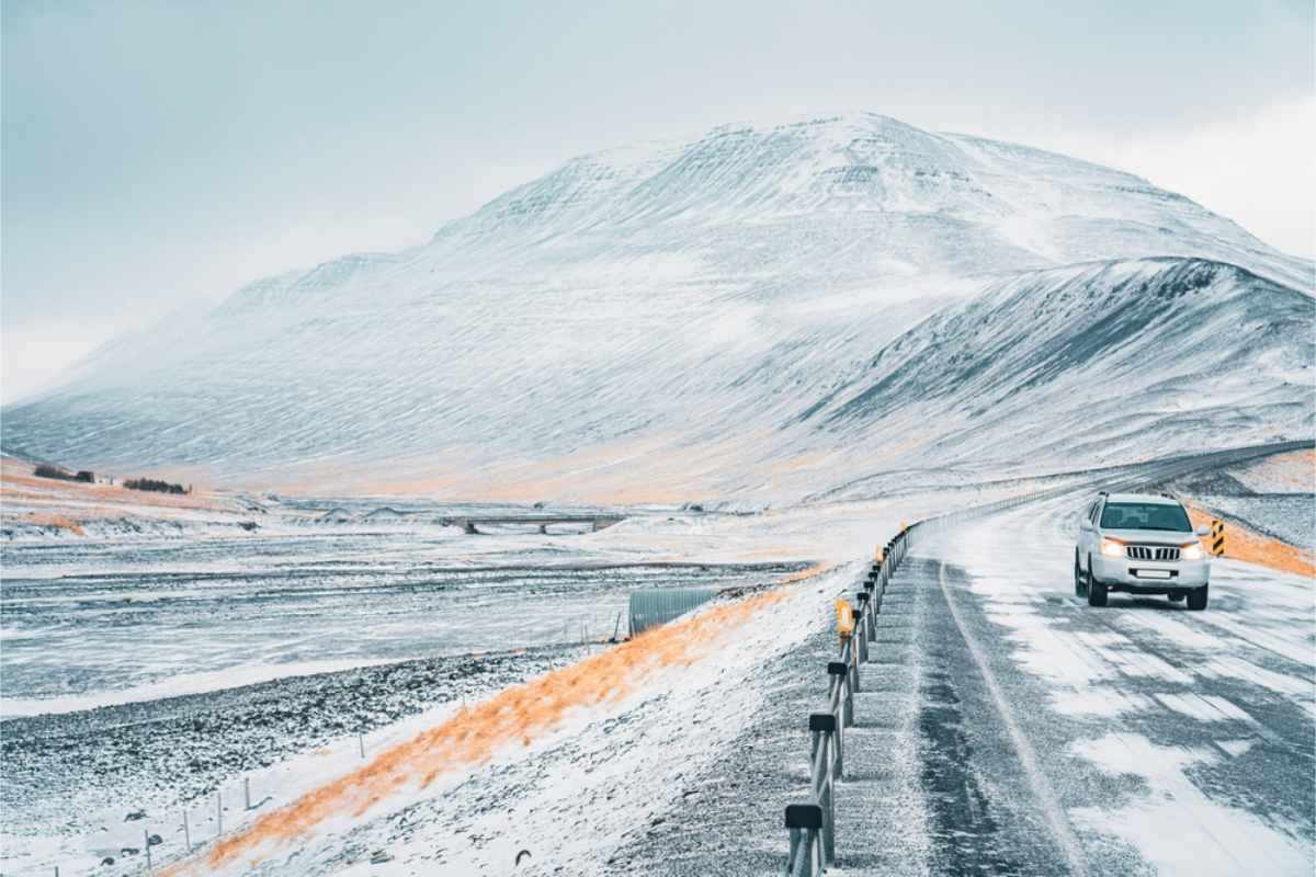 Auto che percorre una strada innevata in Islanda, con montagne coperte di neve sullo sfondo e una luce invernale limpida.